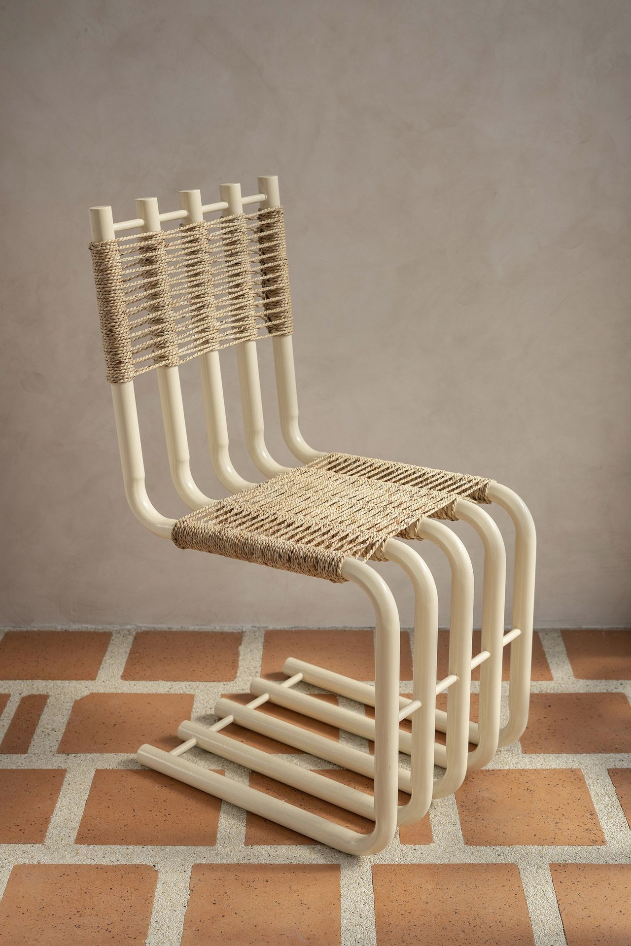 An iron and rattan beige chair stands against a cream background on a brick and cream floor decorated with faux tile motifs.