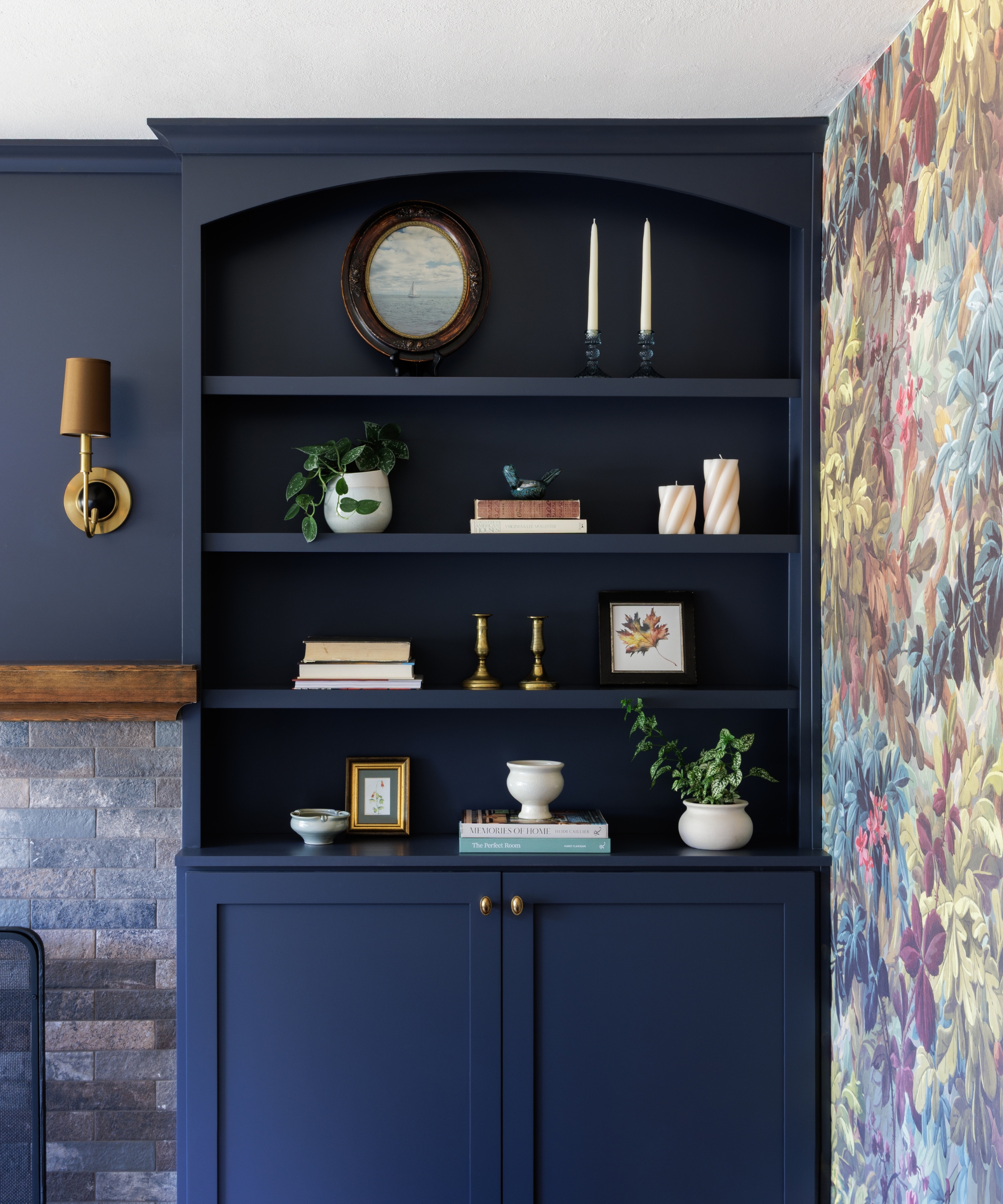 Deep navy built-in shelving with brass hardware, decorative styling, floral wallpaper, and a gray brick fireplace surround.