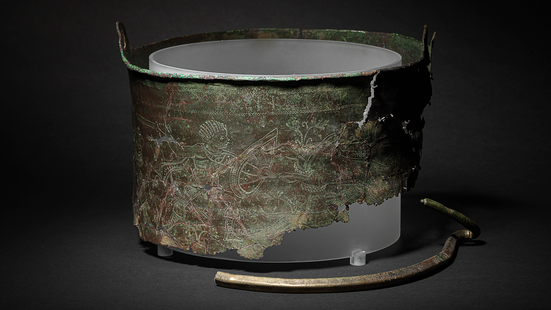 A copper-alloy bucket that has turned brown and green shows incised designs of a person and wild animals