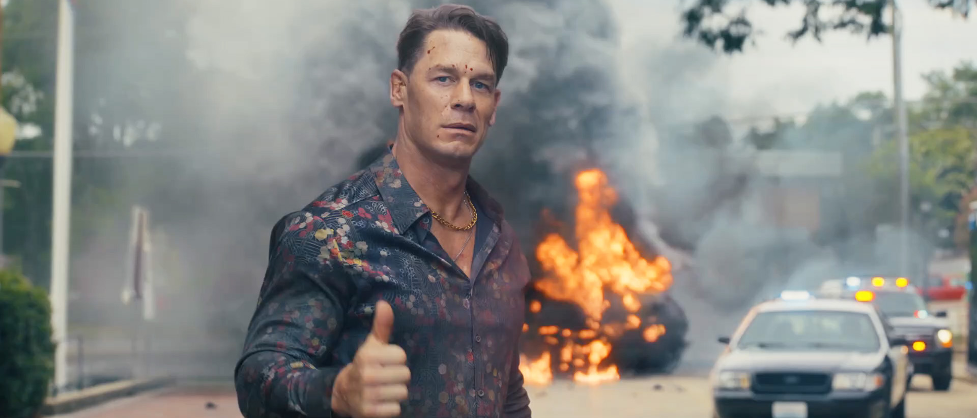 John Cena&#039;s Chris Smith smiling and giving a thumbs up as a car explodes behind him in Peacemaker season 2