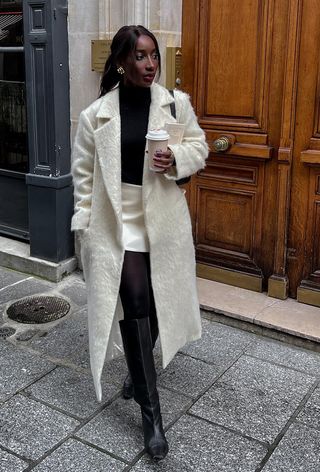 black turtleneck outfit shown in a photo of a French woman on the sidewalk wearing a white mohair coat over a black turtleneck sweater styled with a white mini skirt, black tights, and black knee-high boots