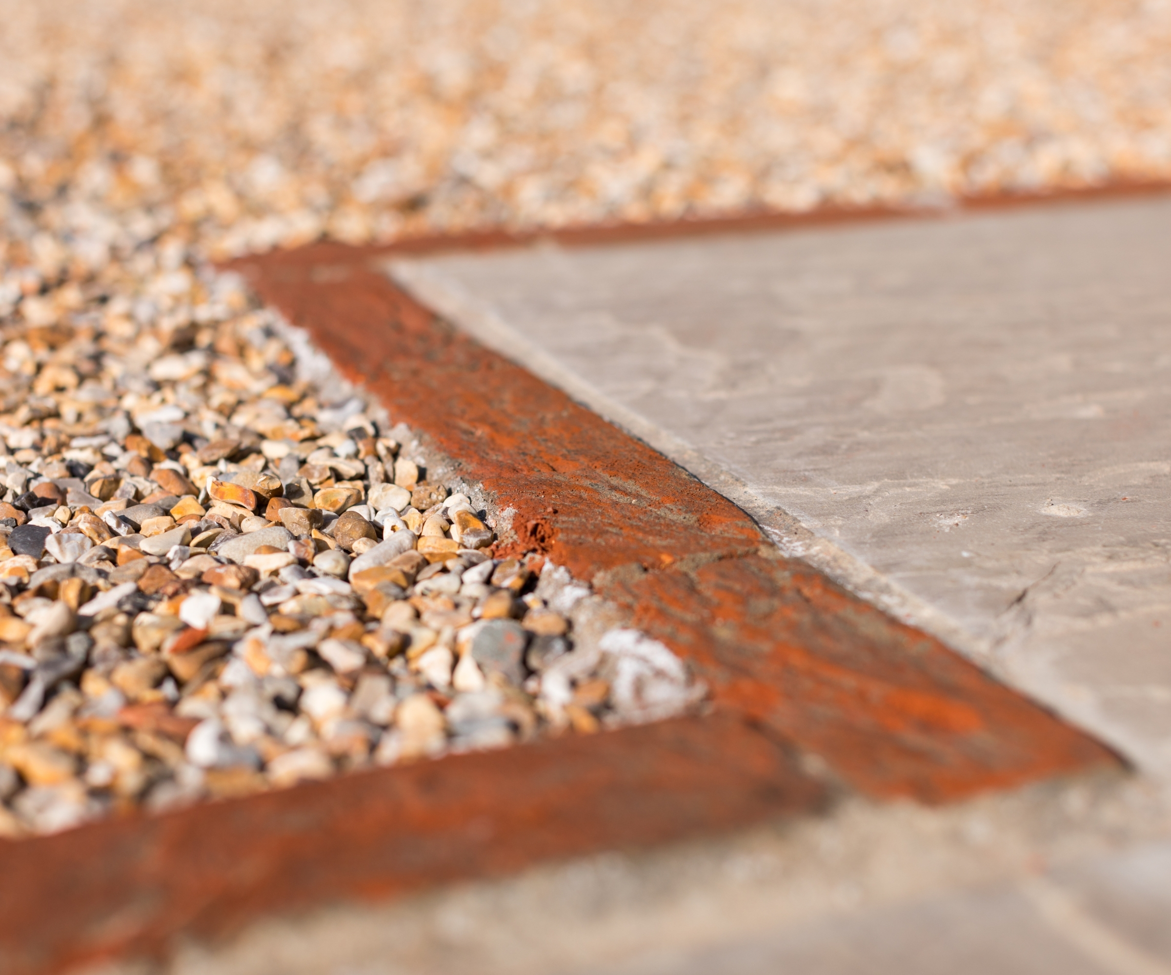 Close-up of a terracotta brick edging strip separating pale paving stones from gravel chippings.