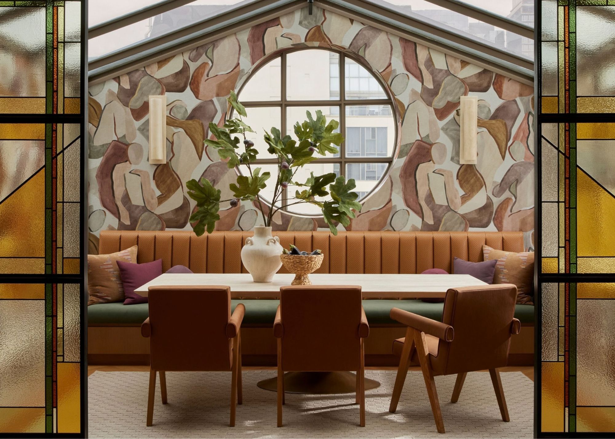 A veranda-style dining room with mural walls, stained glass doors, a pale wood dining table with leather chairs and a brown leather soft bench with green topper, and a thriving plant set in a vase