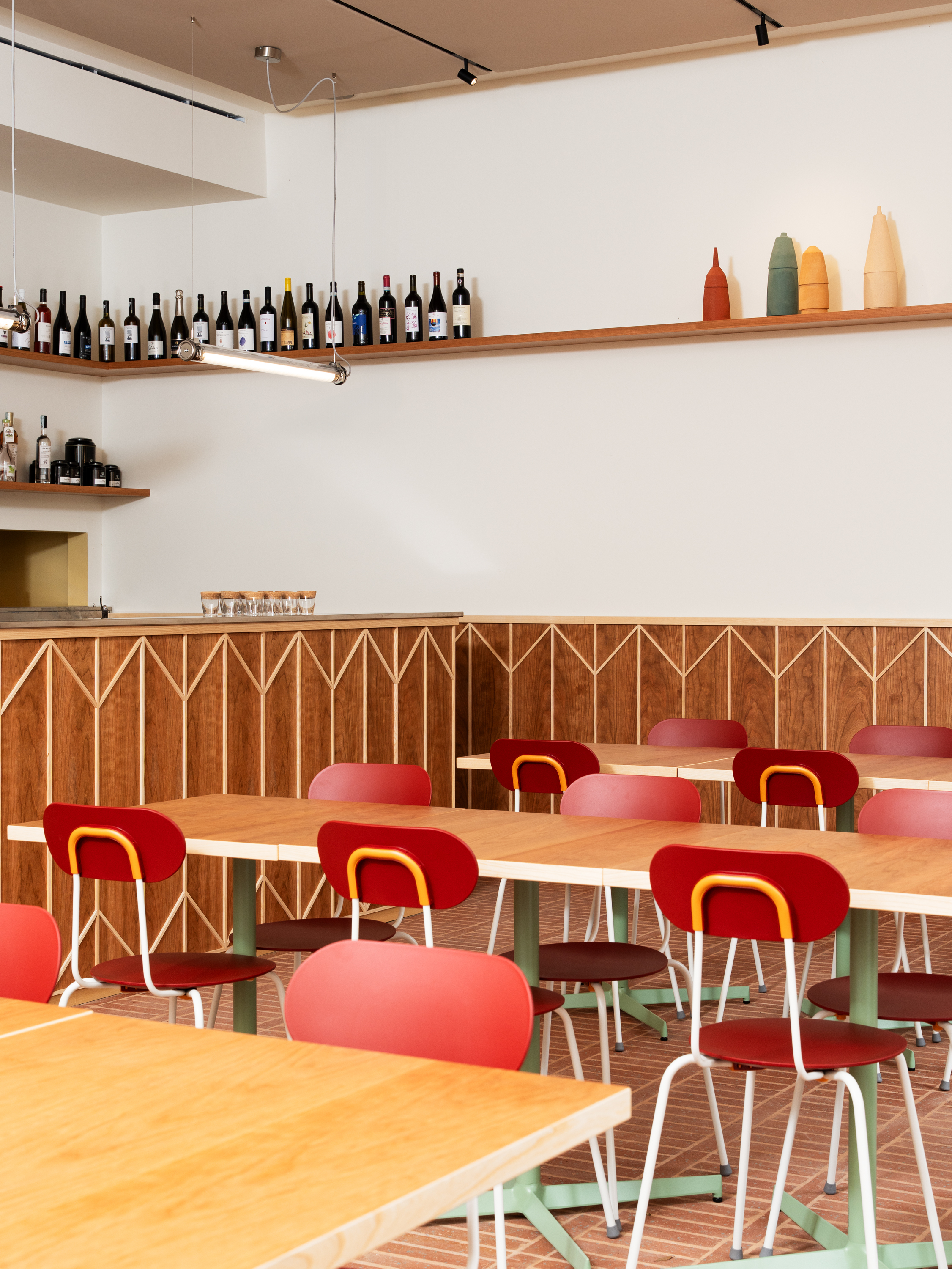 A lively-designed, wood-clad modern osteria features wide wooden tables dotted in bordeaux chairs with white and yellow detailing and decorated with mint green legs.