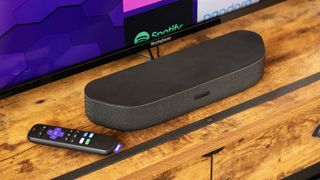 Roku Streambar on a counter top in front of a TV next to a Roku remote