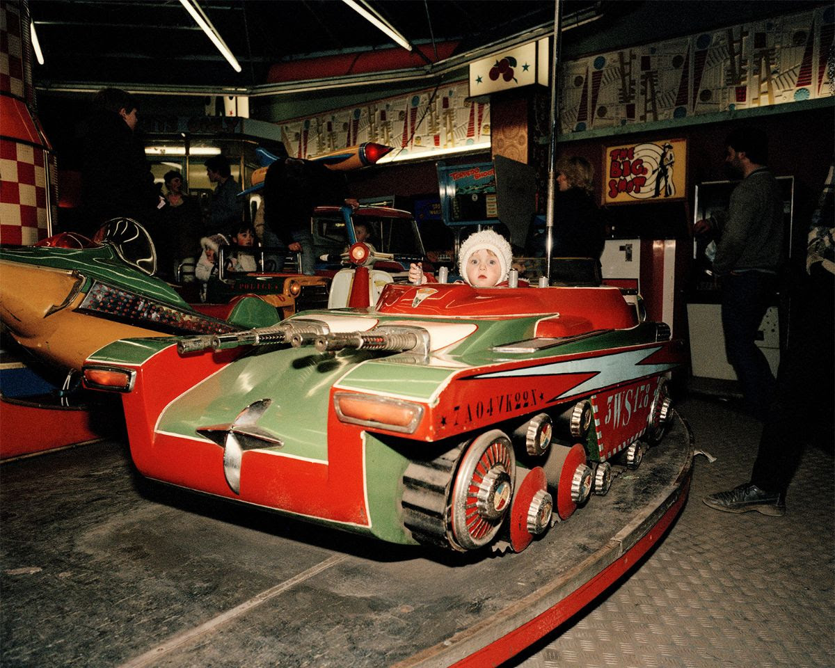 A young child sits inside a colorful, tank-shaped amusement park ride with plastic guns mounted on the front.