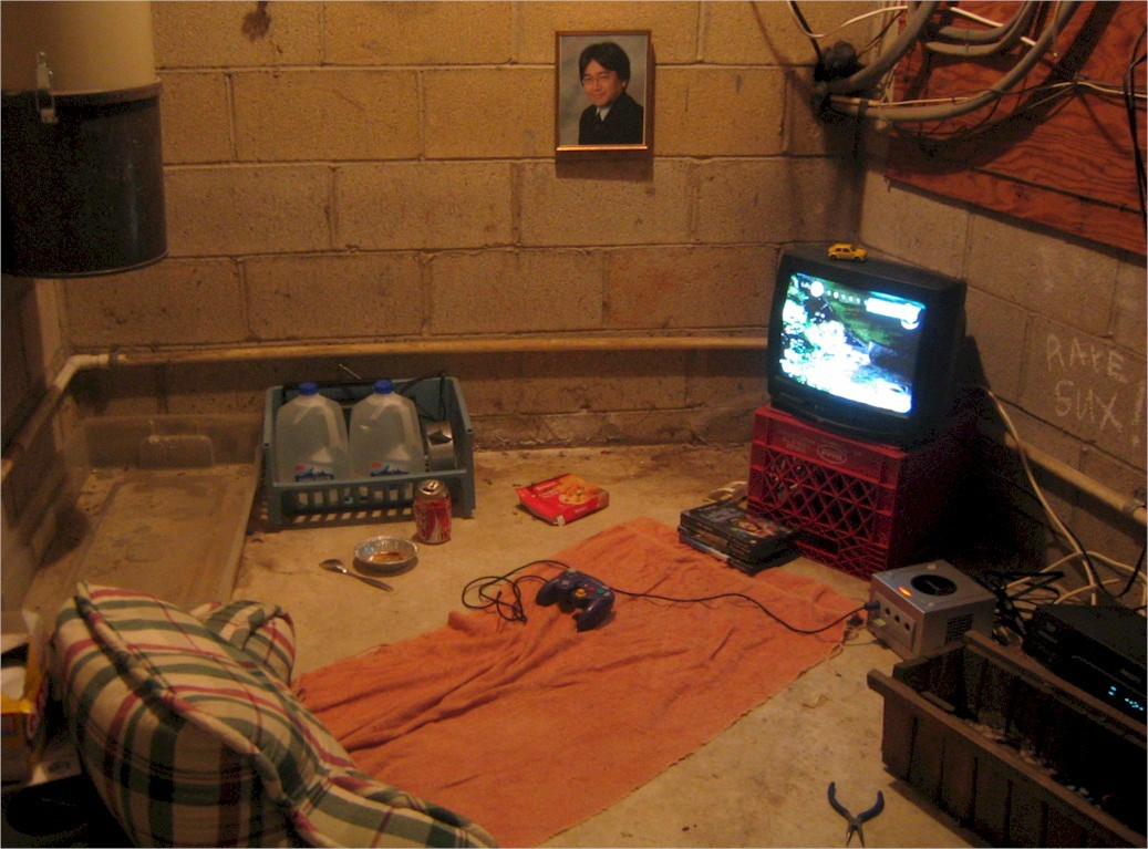 A backjack and towel set on concrete floor in what appears to be a basement, laid out before a CRT TV on a milk crate under the watchful gaze of a portrait of Satoru Iwata.