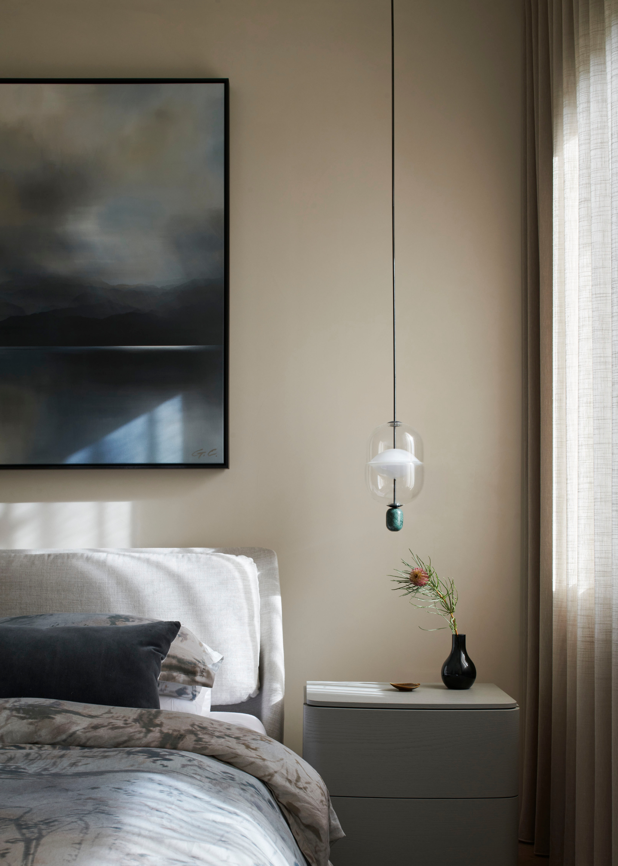 An off-white bedroom with a framed painting above a bed with a patterned duvet and blue pillows, beside a white side table with a vase and a pendant lamp above