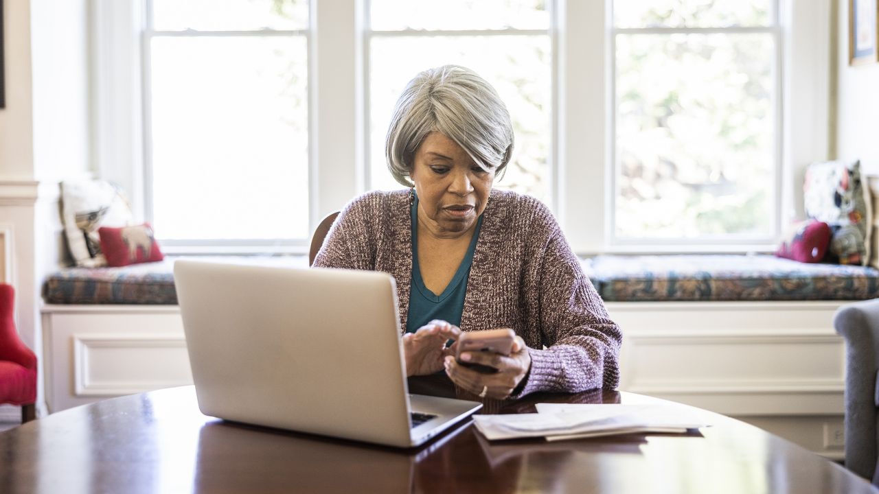 An older woman looks stressed as she looks at her phone while working on her laptop at her dining room table.