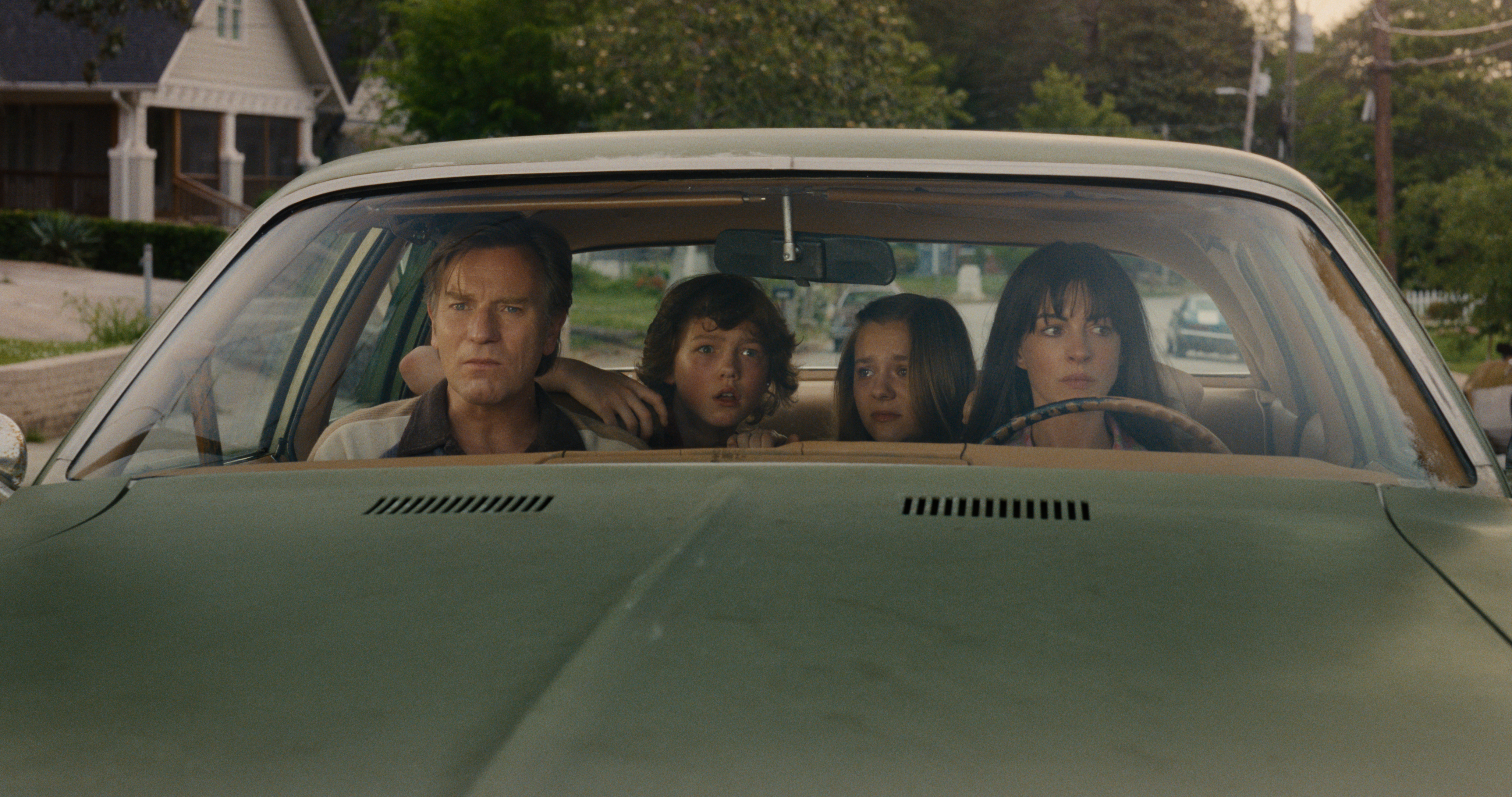 ewan mcgregor and anne hathway as a family driving with their kids in the backseat through a neighborhood in the movie the end of oak street