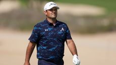 Patrick Reed walks during a DP World Tour event wearing a white cap, white glove and blue/black camo polo shirt