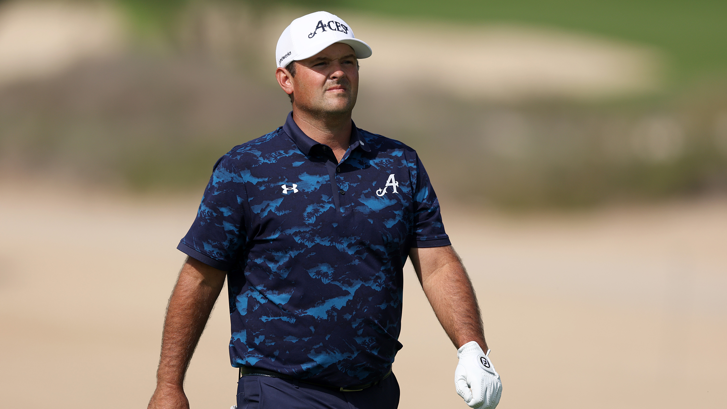 Patrick Reed walks during a DP World Tour event wearing a white cap, white glove and blue/black camo polo shirt