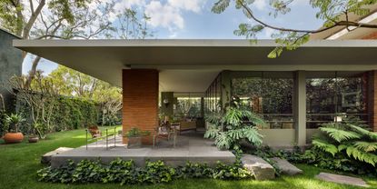 Daytime external image of Ventana House in Mexico, flat white roof, red brick supporting pillar, green lawn, shrubs, plants and trees, windows, stone patio area with irregular shaped stone steps, tables and chairs, blue cloudy sky