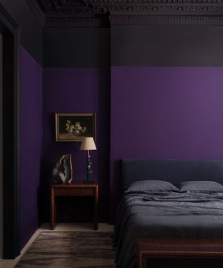 Deep purple bedroom with framed still life, wood side table, intricate stone lamp and ornament and low bed with dark blue linen bedding