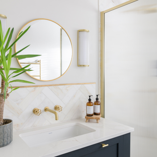 Modern bathoom with white sink and gold taps There is a houseplant and circular mirror.