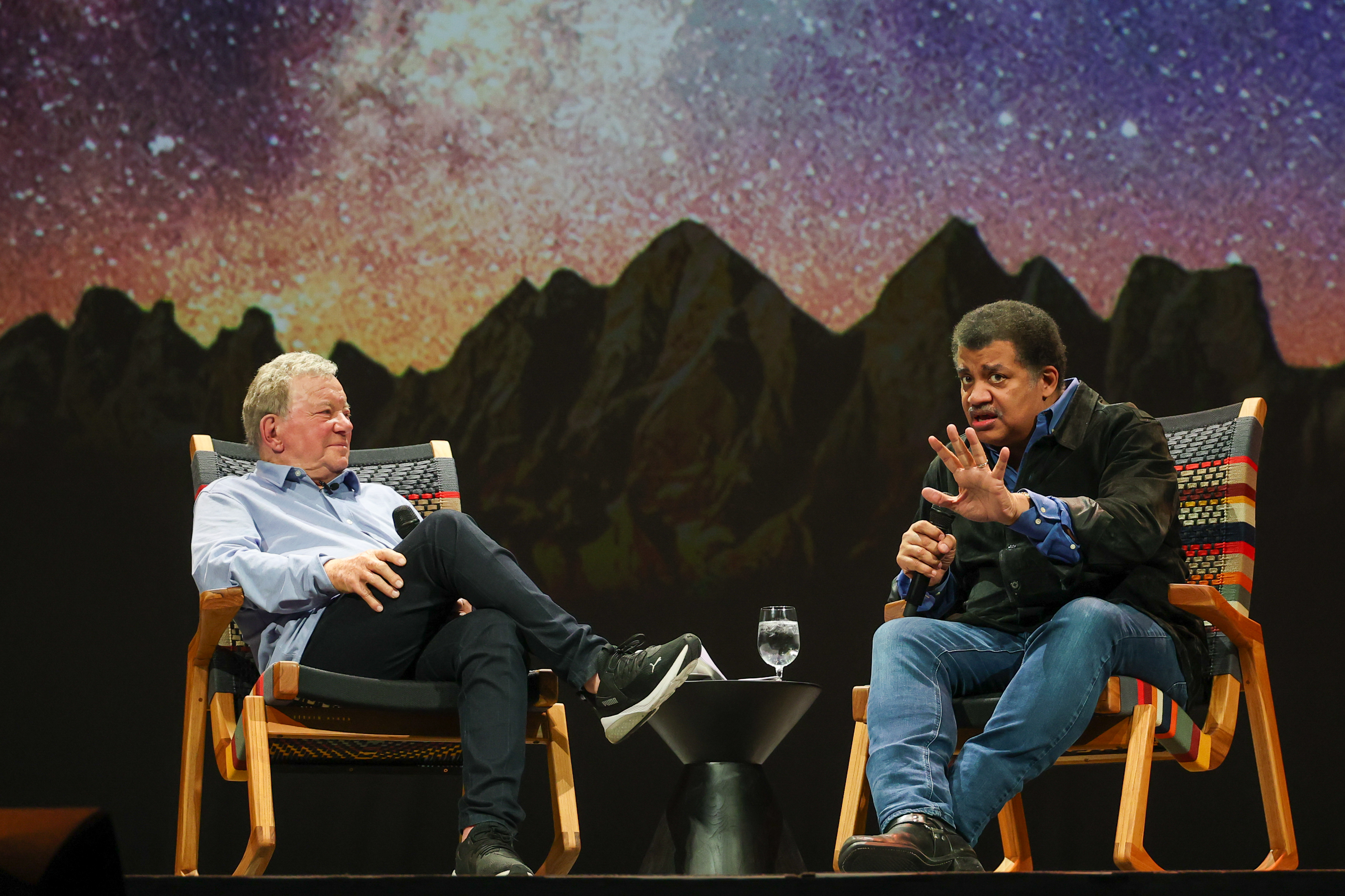 A man wearing a blue shirt stands to the left of a man wearing a black shirt, both in front of a starry background