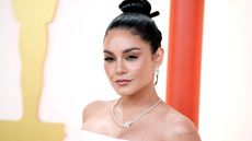 Vanessa Hudgens wearing a white dress, a diamond necklace, and her hair up with a bow, standing in front of a gold and red background