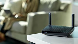 Lifestyle product shot of the Linksys Hydra Pro 6 Wi-Fi router on a side table with a woman on a sofa in the background.