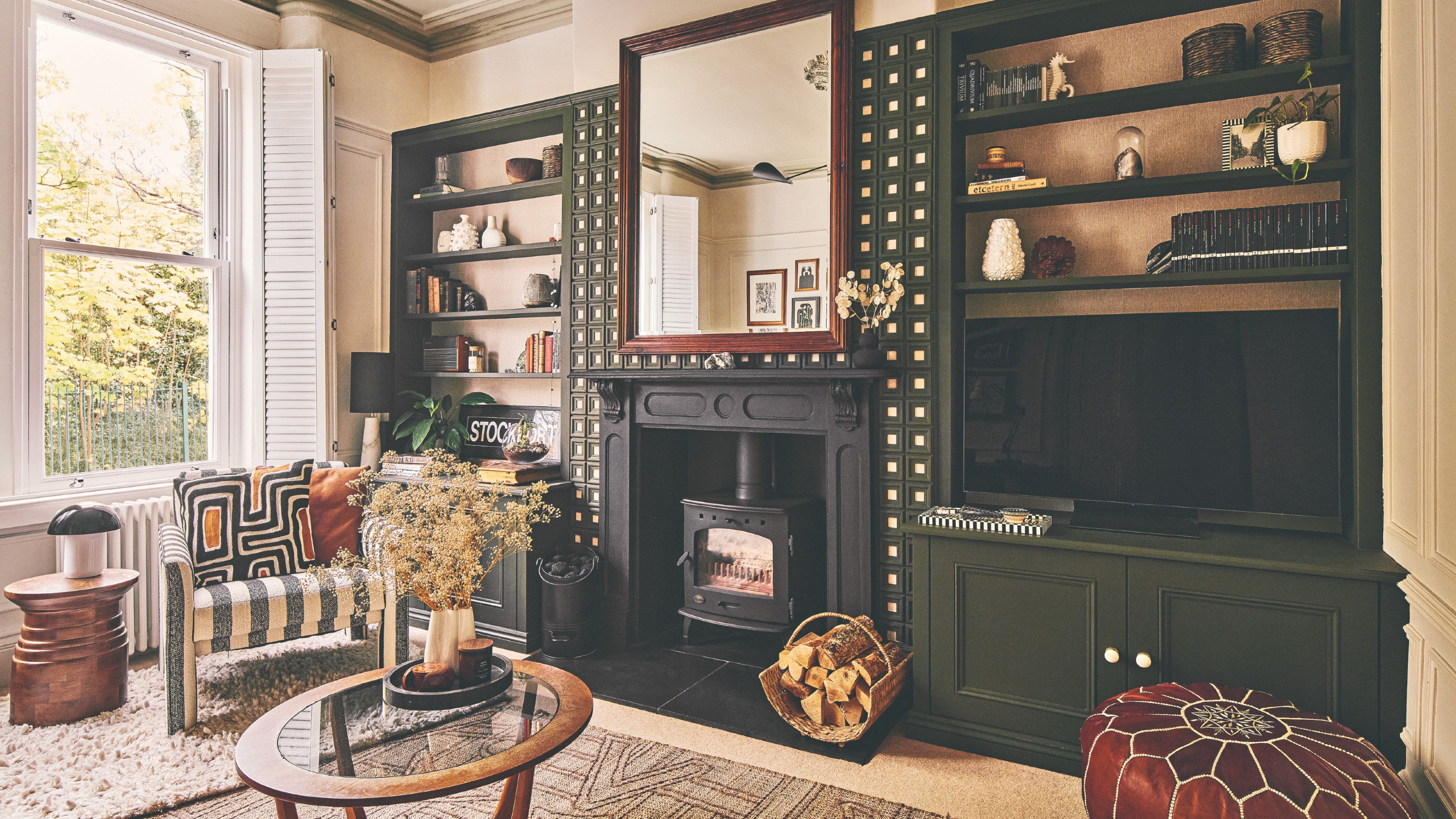 A living room with dark green-painted bookcases fitted into alcoves on either side of the black fireplace with a round glass coffee table and a striped armchair in front of it
