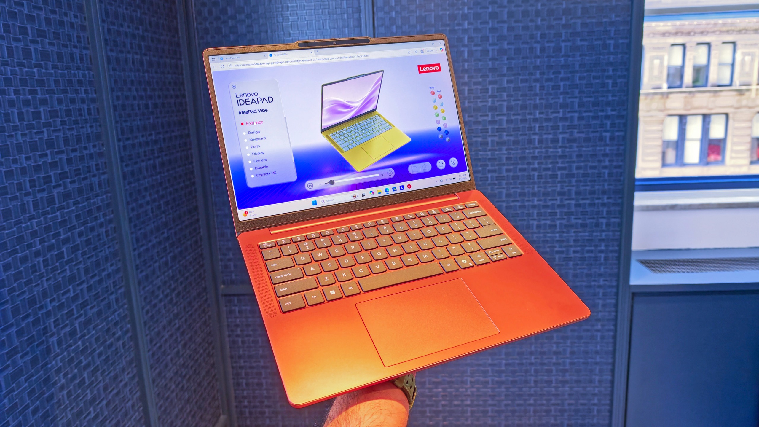 Person holding an open metallic orange Lenovo IdeaPad Vibe laptop displaying its product webpage against a blue textured backdrop.