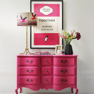 Fuchsia pink colour, with painted French chest of drawers in front of white wall, bamboo table lamp and pink and white poster in black frame.