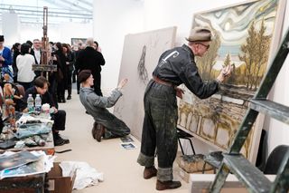 Two painters dressed in vintage workwear give a painting demonstration in a brightly lit gallery filled with people.