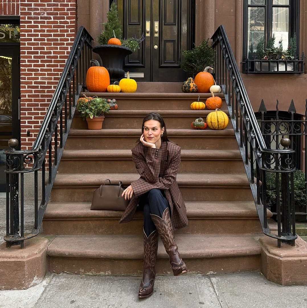 Maggie McCormick wears a a plaid jacket, jeans, and boots in front of a pumpkin-decorated West Village brown stone