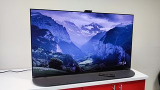 En Sony A95K TV står på en vit TV-bänk med dess tillhörande fjärrkontroller liggandes vid TV-foten. På skärmen visas en bild på snötäckta berg.