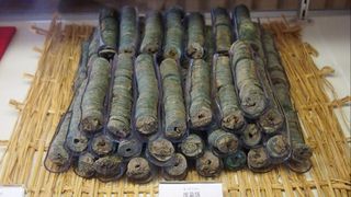 Photograph of stacks of bundled Japanese coins atop a straw woven mat upon a white tabletop.