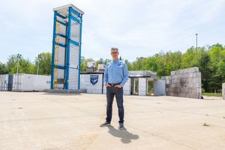 a man in a button-down shirt and jeans stands on a cement tarmac in front of a metal tower