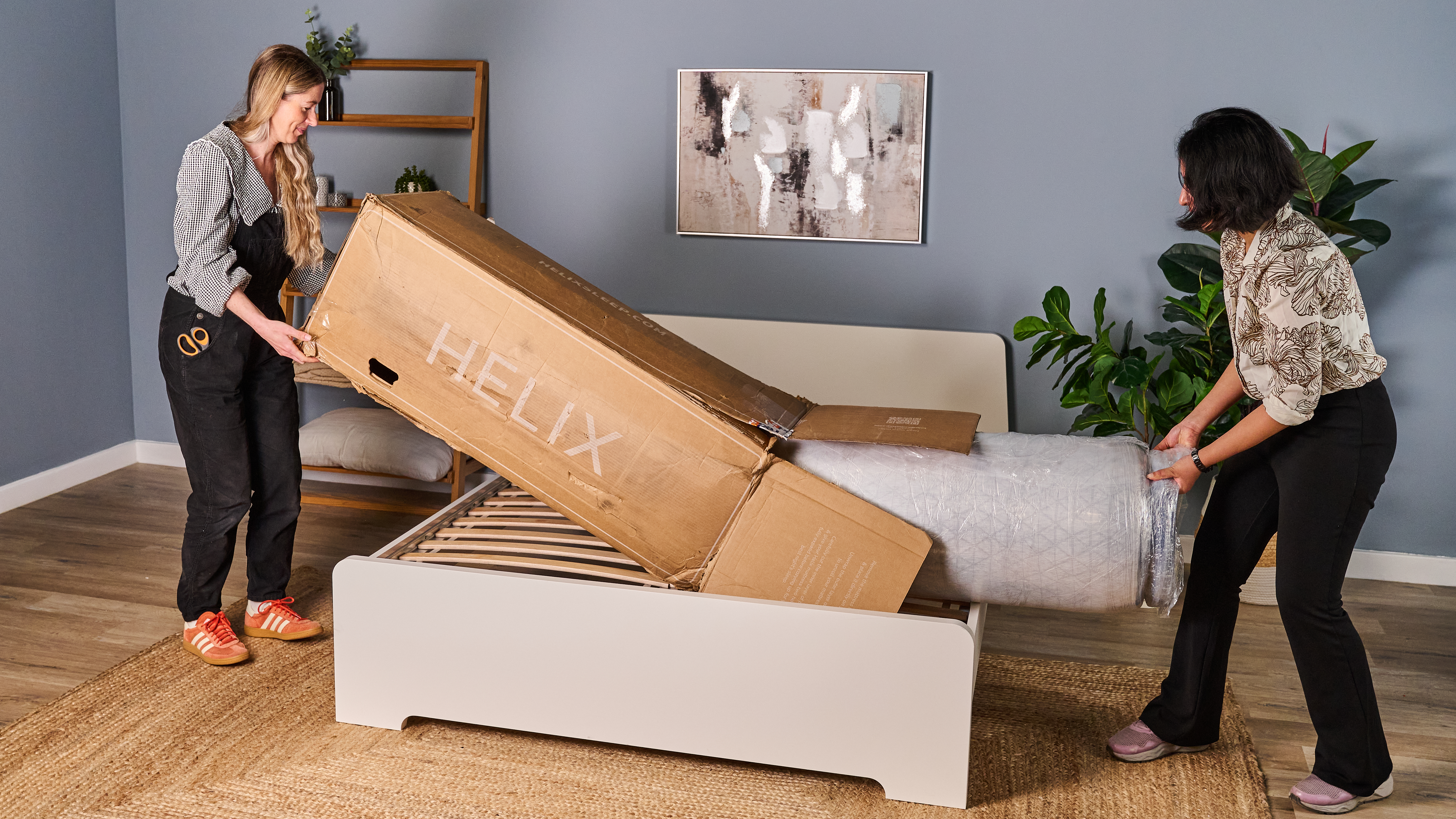 The image shows two mattress testers unboxing the Helix Midnight Luxe mattress and setting it up on a white bed frame in a blue bedroom