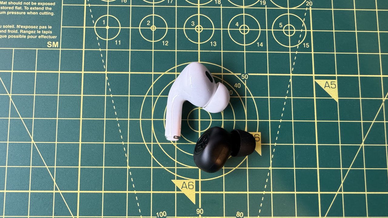 JLab JBuds Mini true wireless headphones on a book and technical mat, with hand and AirPods for comparison.