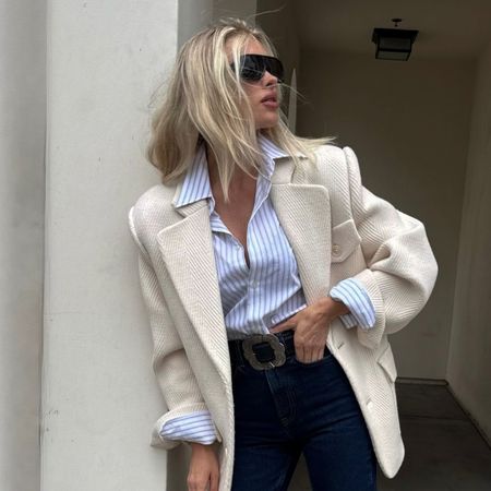 Elsa Hosk poses wearing dark wash cropped jeans with black loafers. She tucked a striped shirt into her jeans and layered a cream blazer on top.