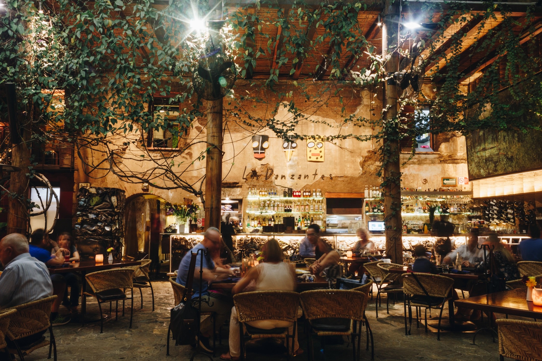 A spirited restaurant featuring modern rustic decor and trailing foliage, along with artworks, a vibrant bar vitrine, and an animated crowd of diners.