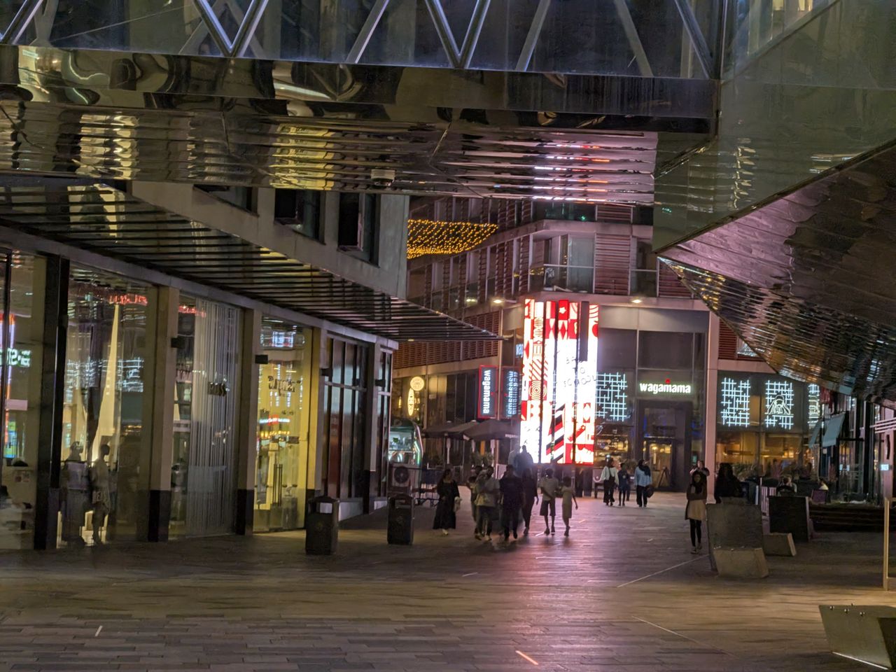 Modern pedestrian shopping street at night, with illuminated glass storefronts and reflective architecture overhead.