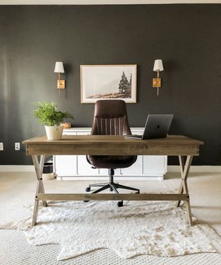 A home office with dark brown walls with two wall sconces and a white wall art print, a brown leather chair, a brown wooden desk with a gray laptop and green plant on it, and a white rug underneath it