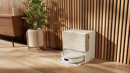 Lifestyle shot of iRobot Roomba 705 Combo in stylish living room on a wooden floor, in front of a wooden-paneled wall. A potted indoor tree and drawers are pictured to the left of the white robot vacuum docked in its cleaning station.