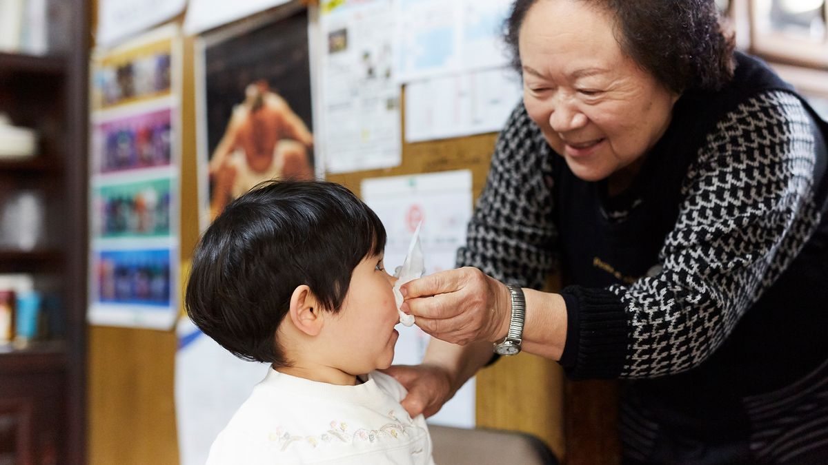 a grandmother smiles as she pinches a tissue over her young grandchild&#039;s nose; both are standing in what looks like a store with posters on the walls