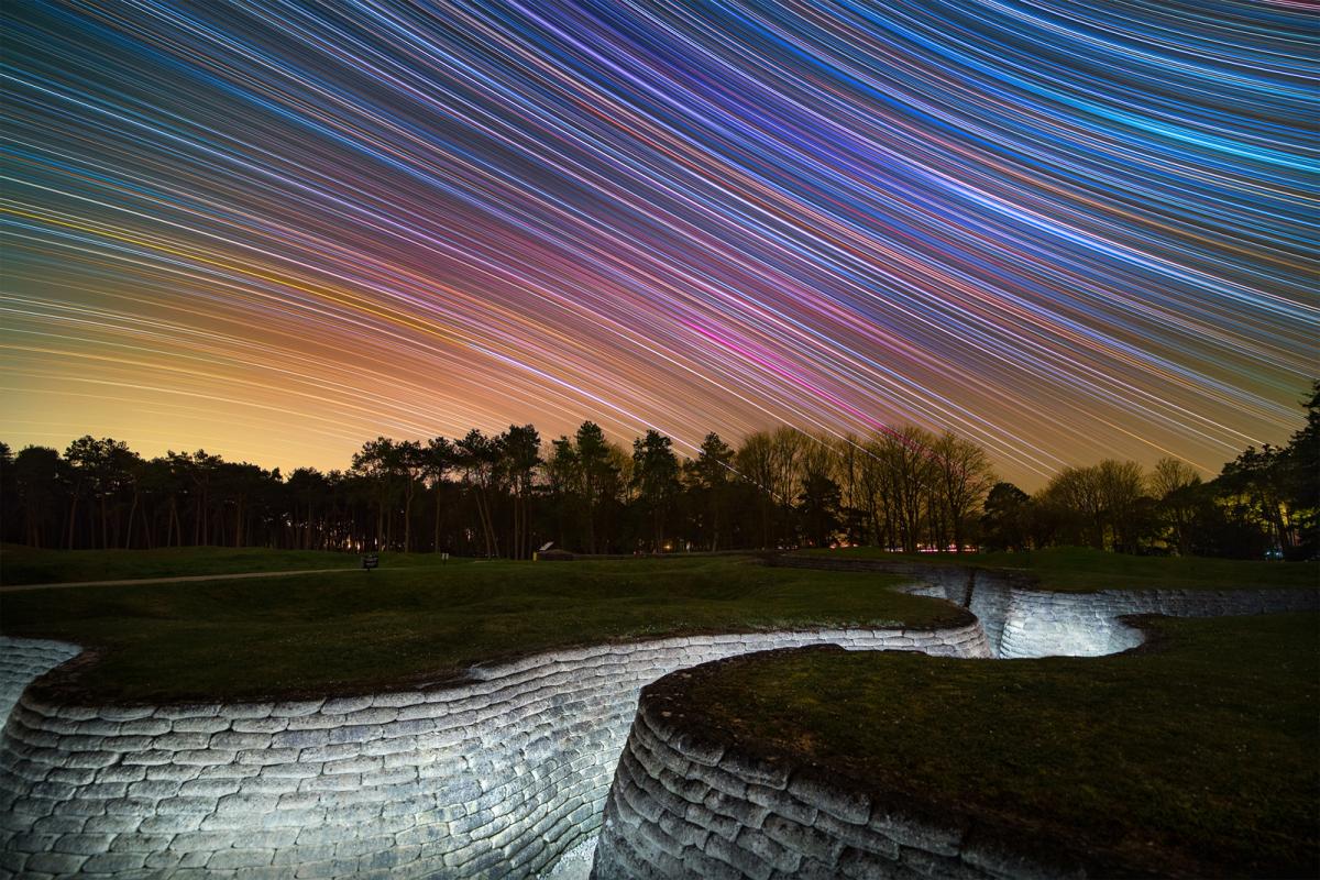 A white-bricked trench lays recessed in the pale green grass of a World War I memorial in France. Above, the night sky is diagonally streaked with a rainbow of stars.