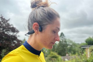 This image shows a woman wearing the Haylou PurFree headphones over her right ear. She has her hair tied up and has a yellow jersey on. There is greenery and a grey sky in the background