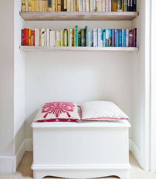 a reading nook on a landing with a white bench seat with pink and white cushions underneath a white bookshelf containing a selection of colourful books