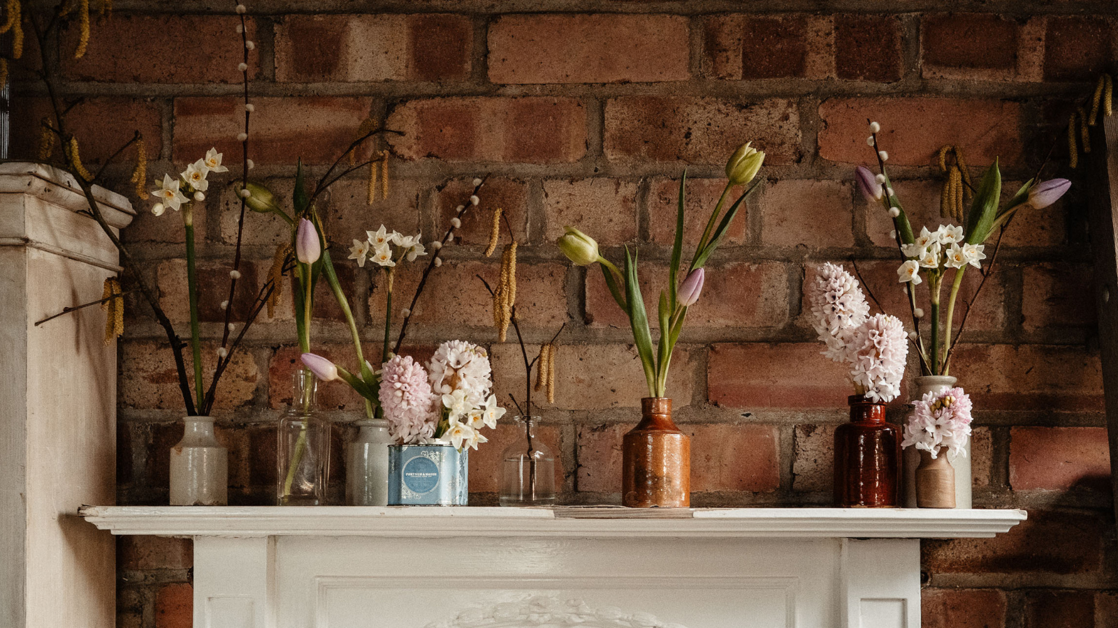 Floral design in vintage vessels and ceramic ink wells on the mantelpiece against red brick wall background 