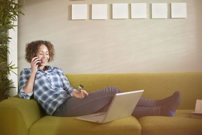 a woman sits on a sofa with her laptop and credit card and chats on the phone