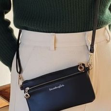 Woman wearing green jumper, white pants, and black Save the Girls leather crossbody phone bag.