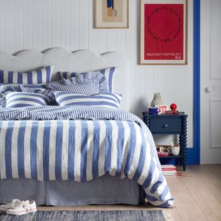 Blue and white striped bed linen on a double bed with a scalloped headboard in a white bedroom with painted tongue and groove walls