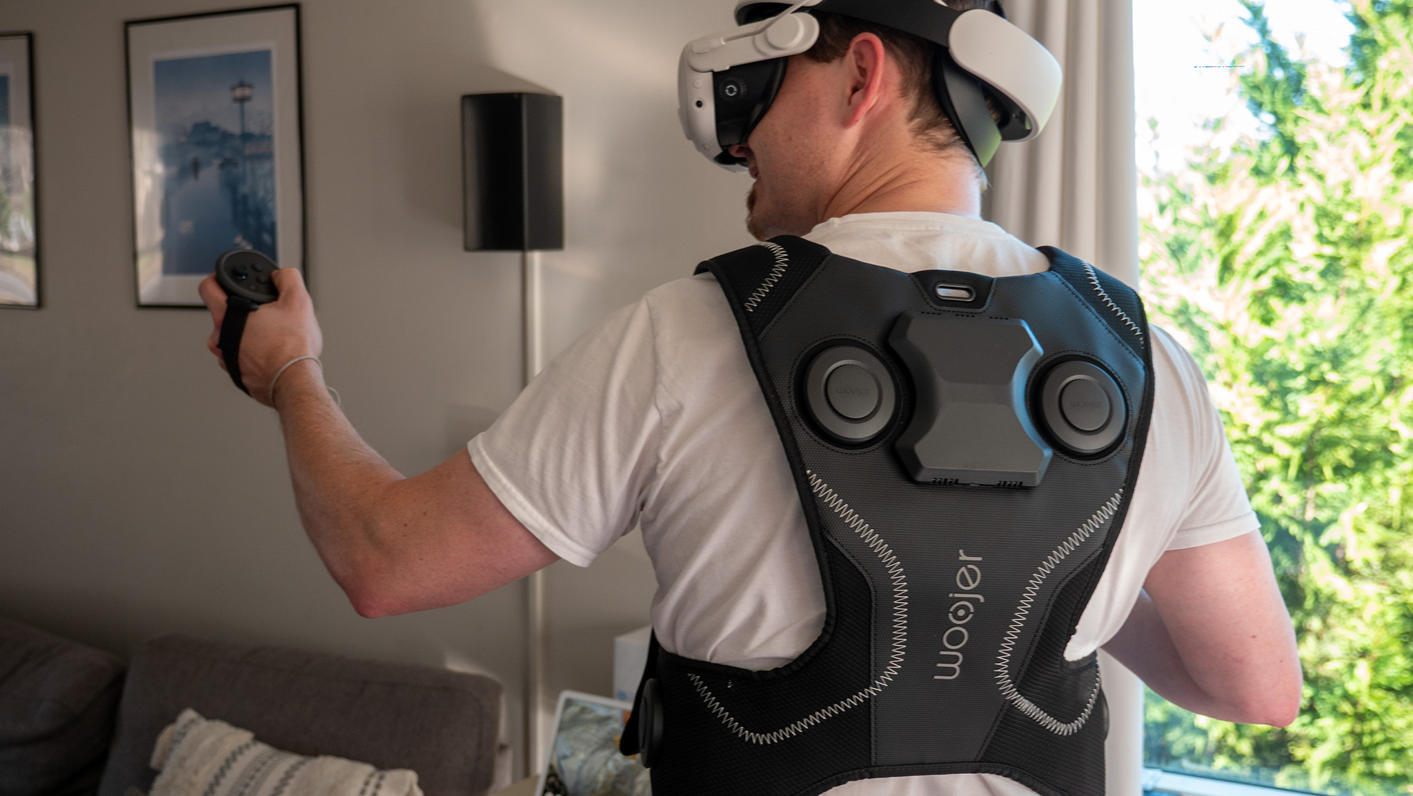 A photo of a man wearing the Meta Quest 3 and the Woojer Haptic Vest 4 from the back, holding up a controller.