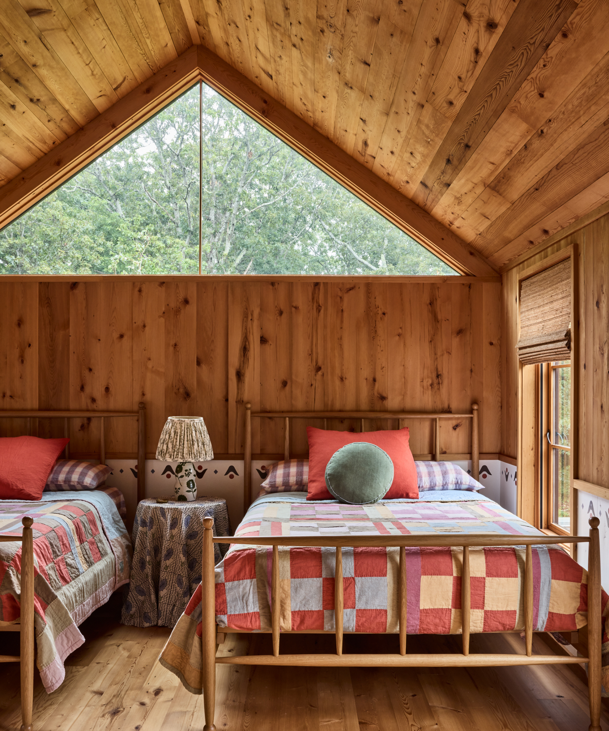 Rustic twin bedroom with wood paneling, sloped ceiling, an A-frame window overlooking trees, and bright patchwork quilts