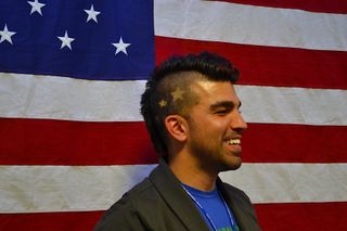 A man wearing a mohawk haircut stands before an American flag