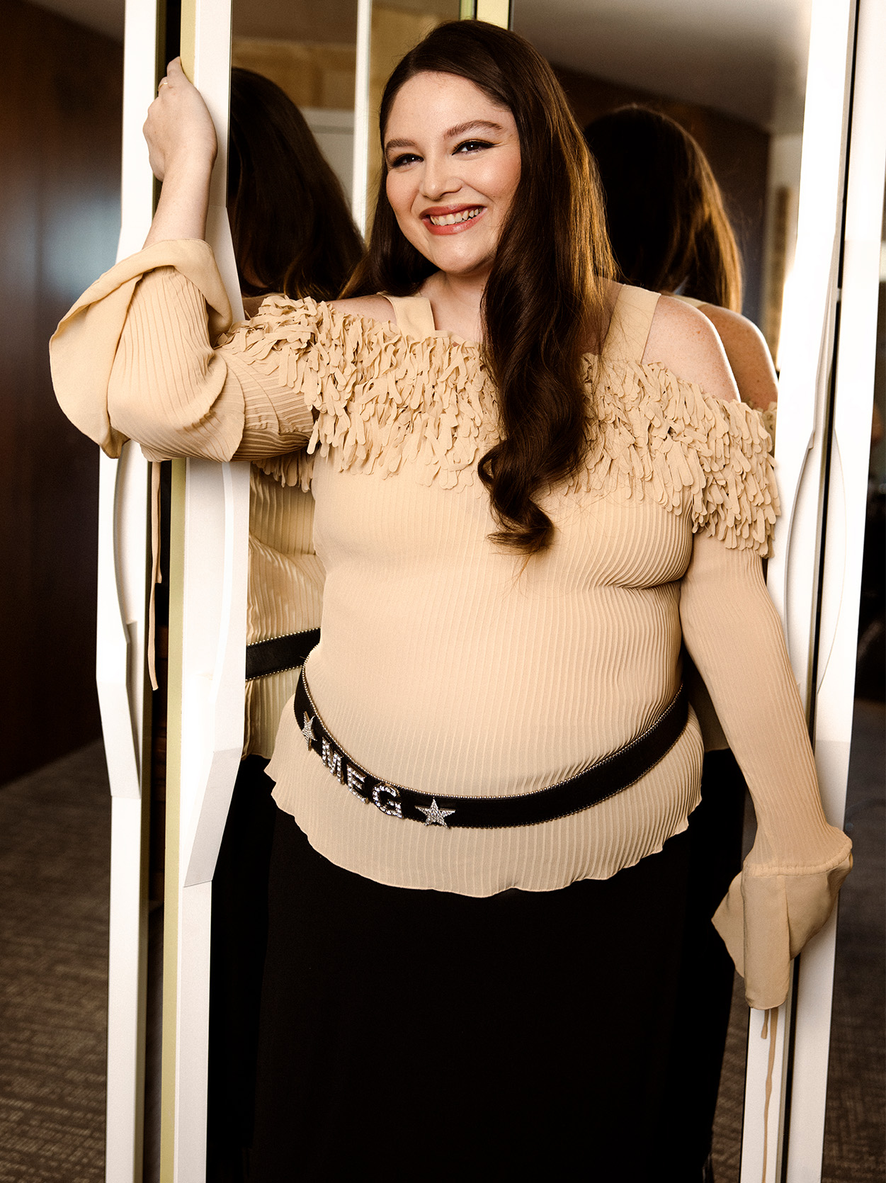 actress Meg Stalter smiles posing against a mirror wearing a biege off the shoulder sweater black skirt and a belt that says meg