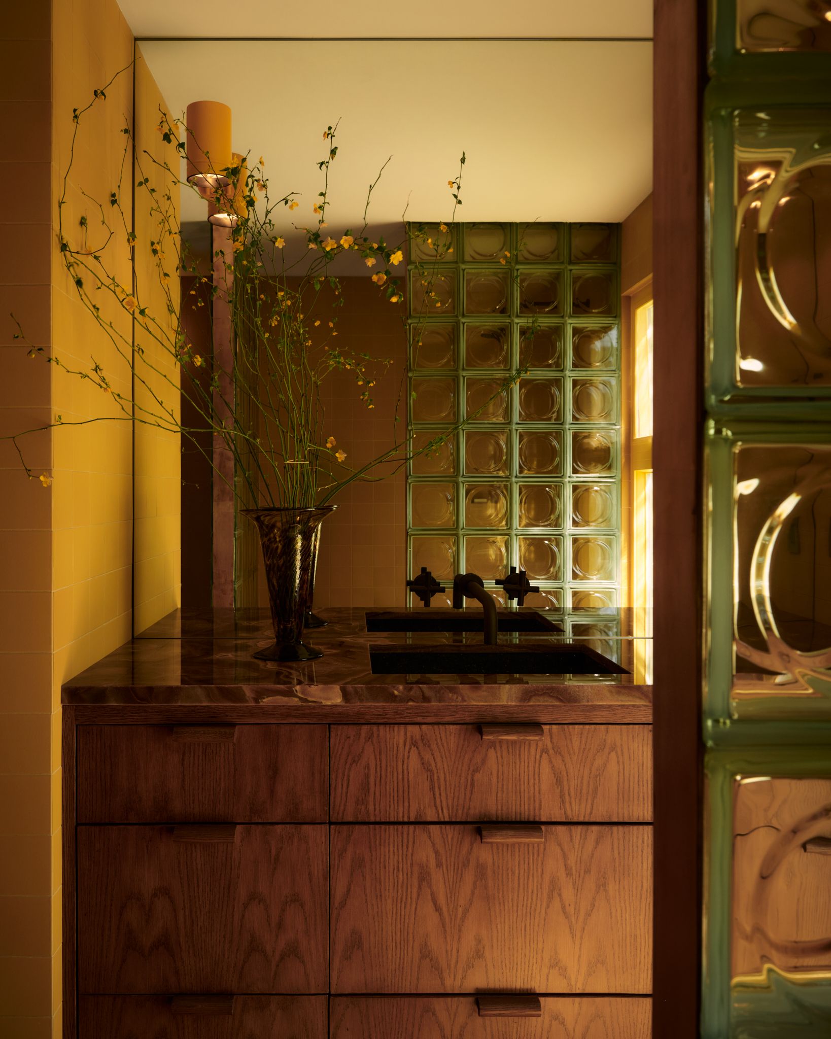 A modern bathroom with a large mirror over the wooden vanity with green-framed glass blocks reflecting in the mirror and a vase of long-stemmed thin flowers by the sink